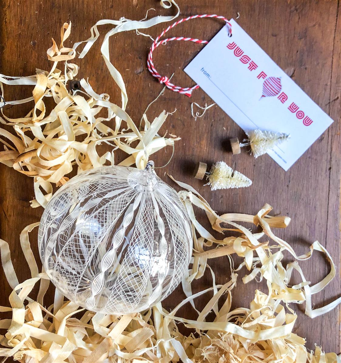 2020 Ballotini ornament, combined with a THF-designed gift tag clear and white glass ornament, sitting on table with excelsior, tiny Christmas trees, and a gift tag