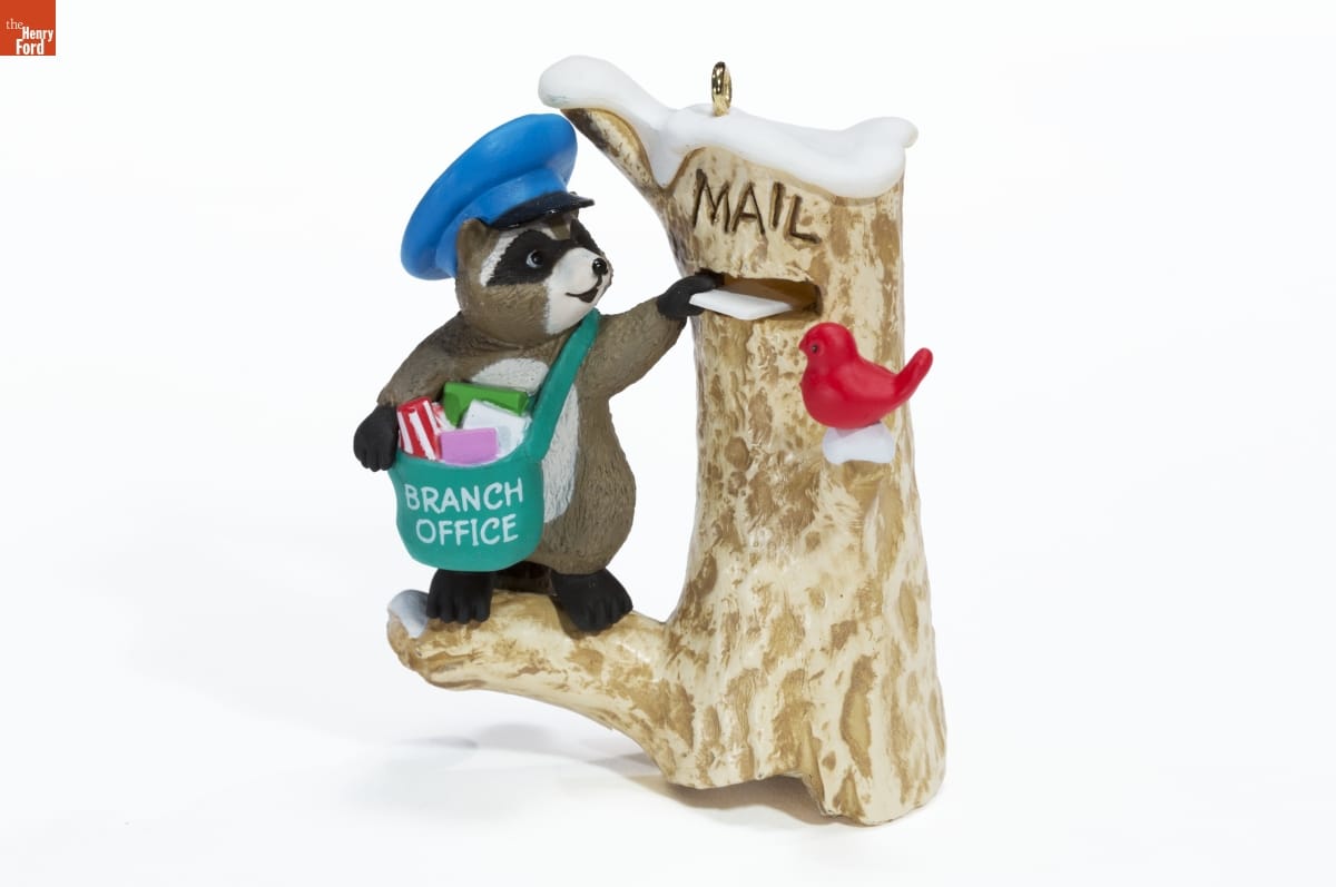 Figurine of raccoon wearing cap and mailbag putting a letter into a slot in a tree trunk