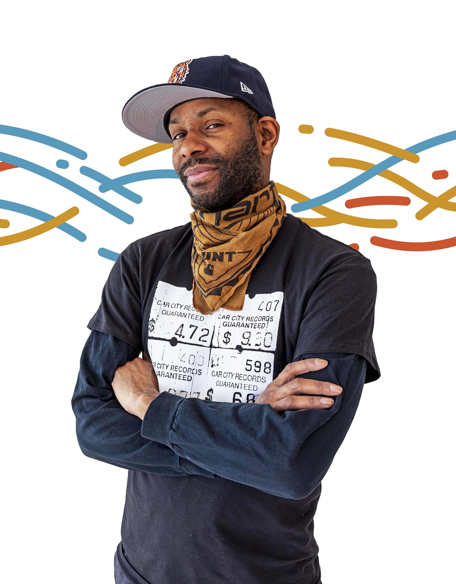 Waajeed, director of the Underground Music Academy African American man wearing long-sleeve t-shirt, bandana around neck, and baseball cap stands with arms crossed with a colorful pattern of wavy lines in the background