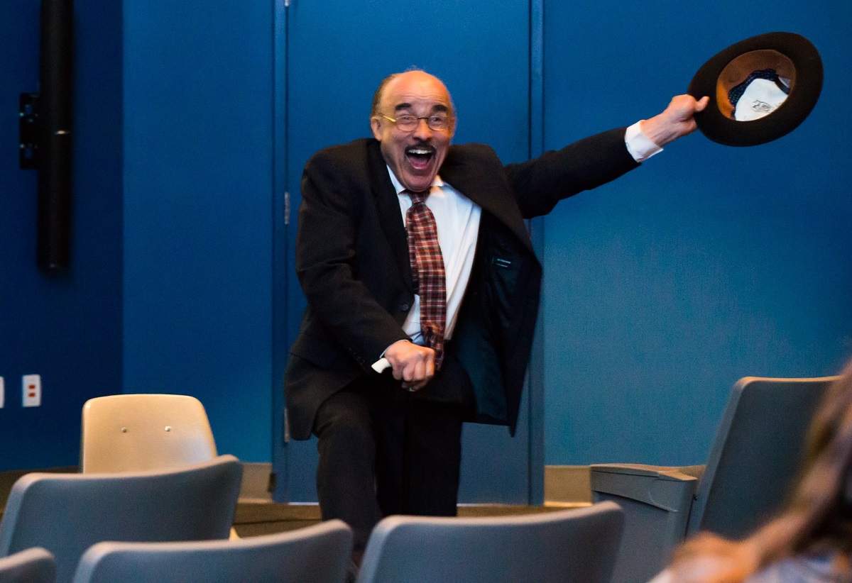 Man in suit smiles and holds a hat to the side among chairs