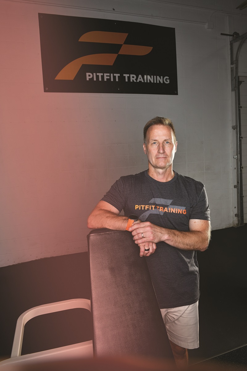 Man in t-shirt and shorts stands under a sign reading "PitFit Training"