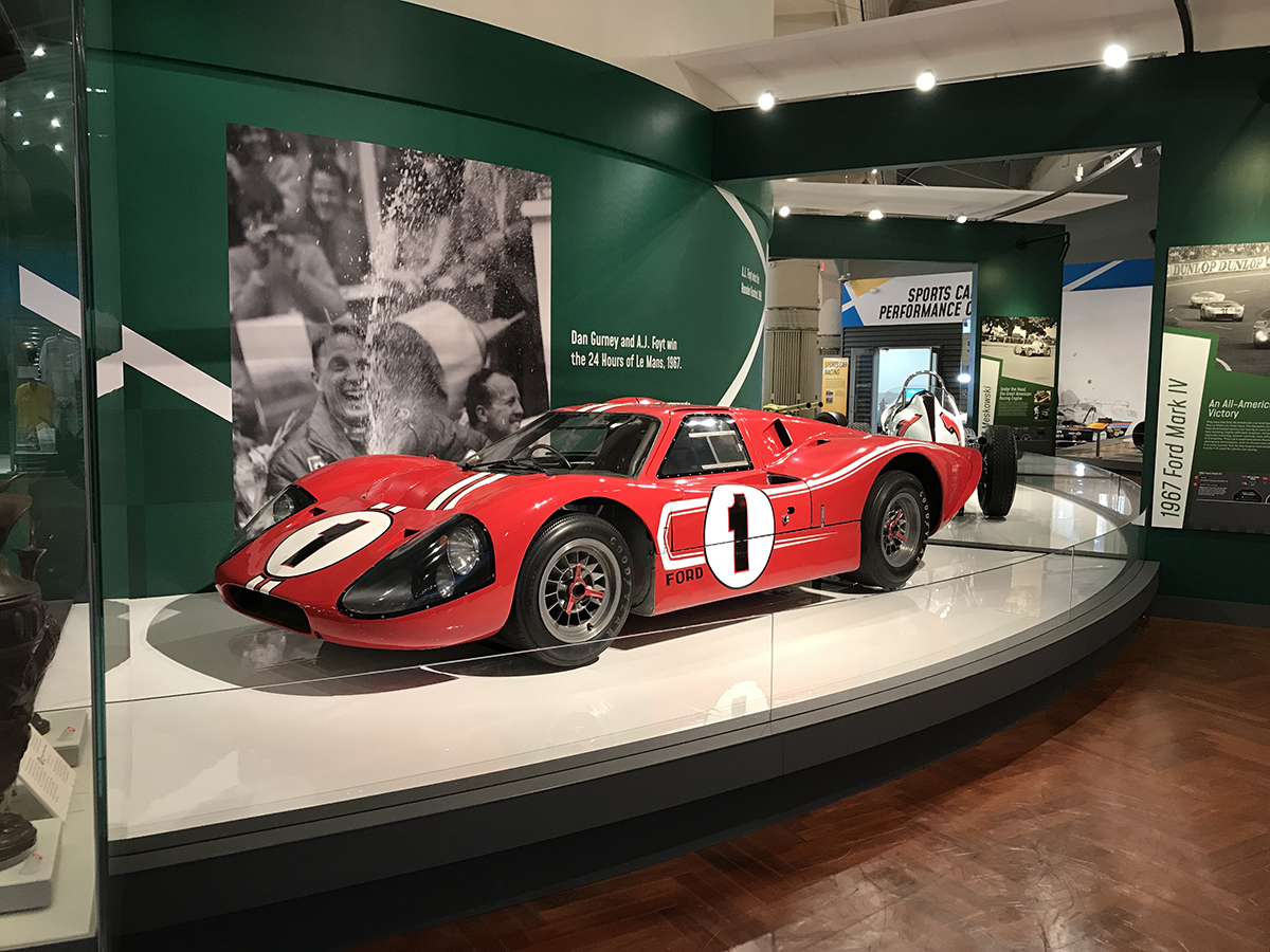 The Mark IV on permanent display in Driven to Win. Low red race car with number "1" in a circle on side sits on a platform in front of a green backdrop with a large photo and text