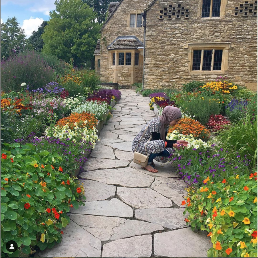 Woman in a dress and hijab kneels on a gravel path through a flowerbed with a beige stone cottage in the background
