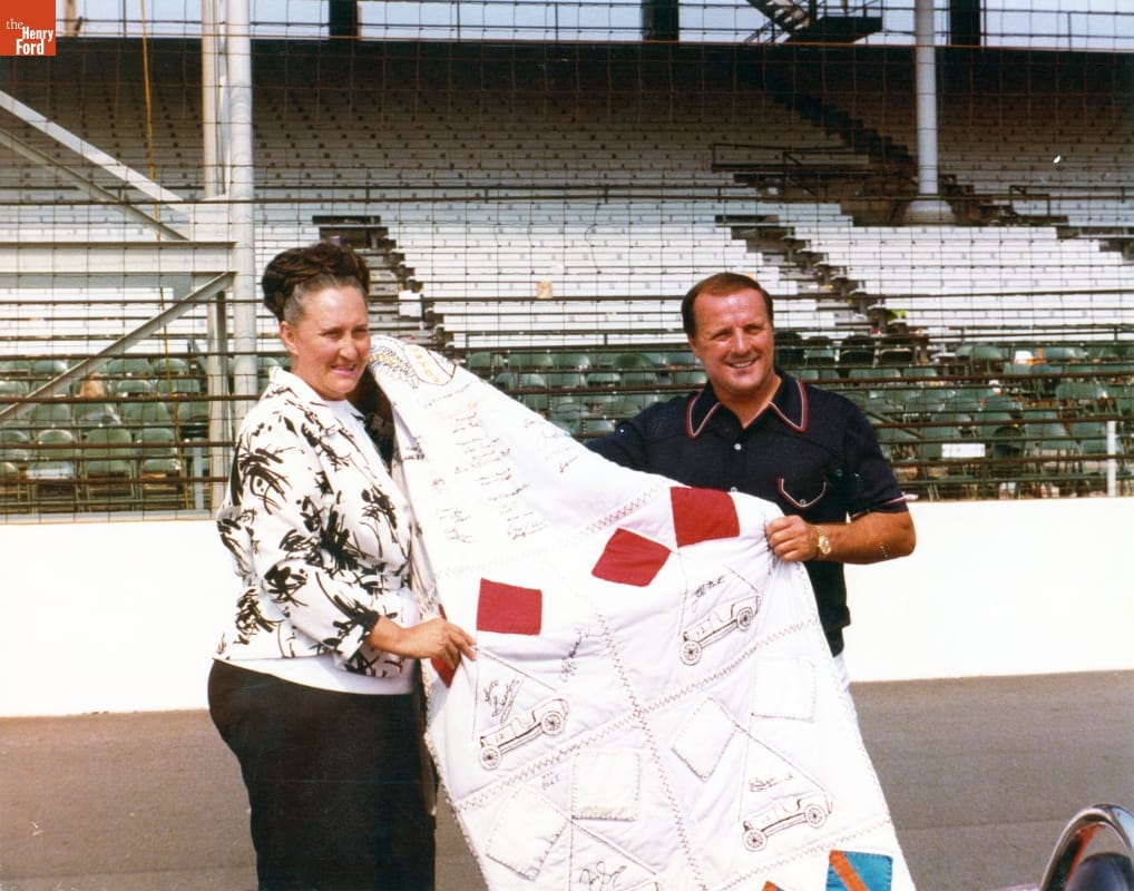 Woman with dark hair in braid wrapped around her head and man in polo shirt hold a quilt in front of empty grandstands