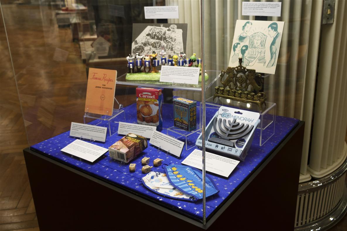 Hanukkah case in Henry Ford Museum of American Innovation Display case with dark base and clear cover, containing various items on lucite display stands and cards with text on them