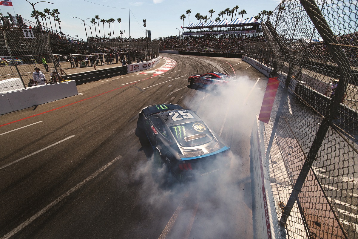 Vaughn Gittin, Jr., during the 2019 Formula Drift championship series Two race cars drift and spew smoke on a racetrack surrounded by fencing, palm trees, and spectators in stands