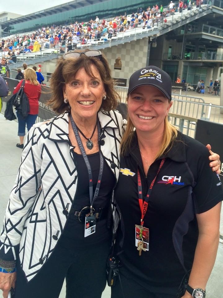 Sarah Fisher with Lyn St. James Two women wearing lanyards pose with arms around each other, with a grandstand full of people in the background