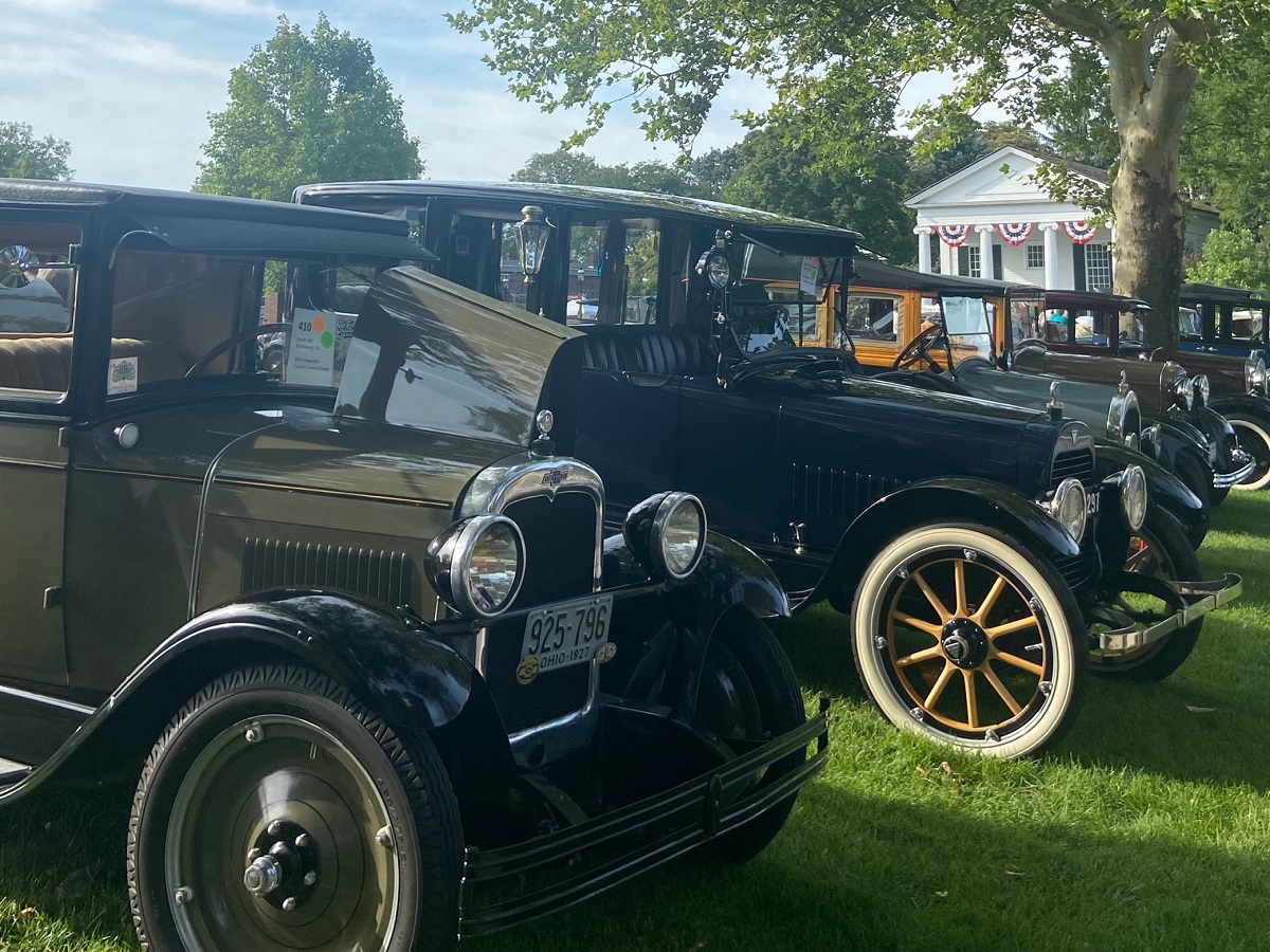 Vehicles participating in Old Car Festival 2022 on the Village Green A row of old-fashioned cars are parked on a green lawn in front of a white building with columns decorated with red, white, and blue bunting