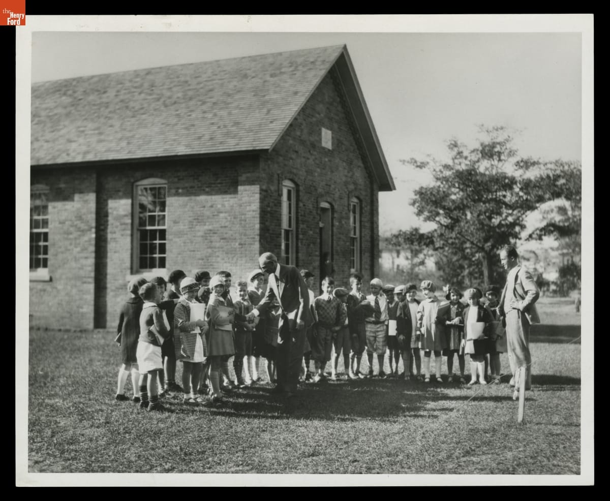 Two men stand with a group of children outside a small brick building; one shakes hands with a child