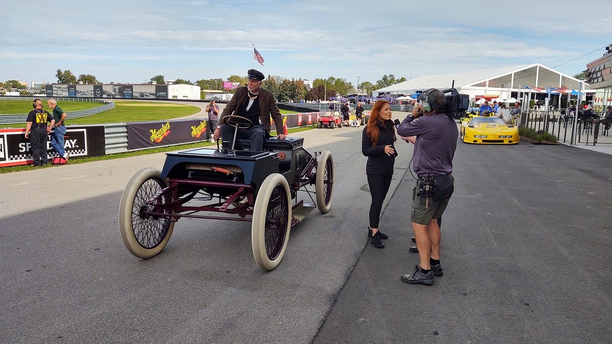 “Sweepstakes” replica at the 2021 American Speed Festival Man sits in boxy open early car on racetrack; a woman stands nearby being filmed by a cameraman