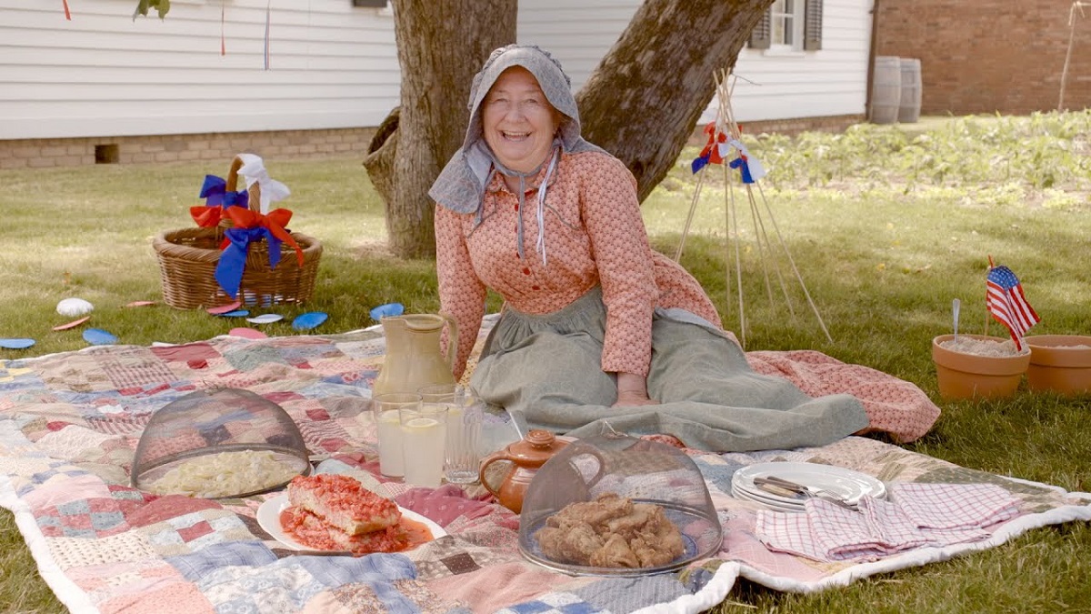 Woman in gingham dress, bonnet, and apron sits on a picnic blanket with food in front of a tree and white wooden house 