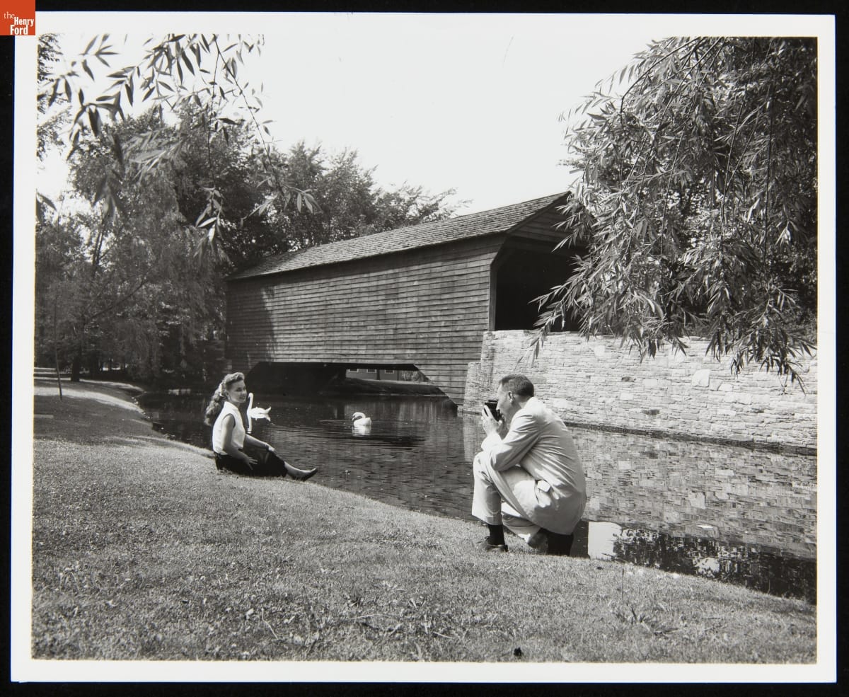 Lloyd Van Meter Photographing Nancy Lawrence near Ackley Covered Bridge in Greenfield Village, June 1958 Black-and-white photograph of man, kneeling, photographing woman seated on grass, with pond, swans, and a wooden covered bridge behind them
