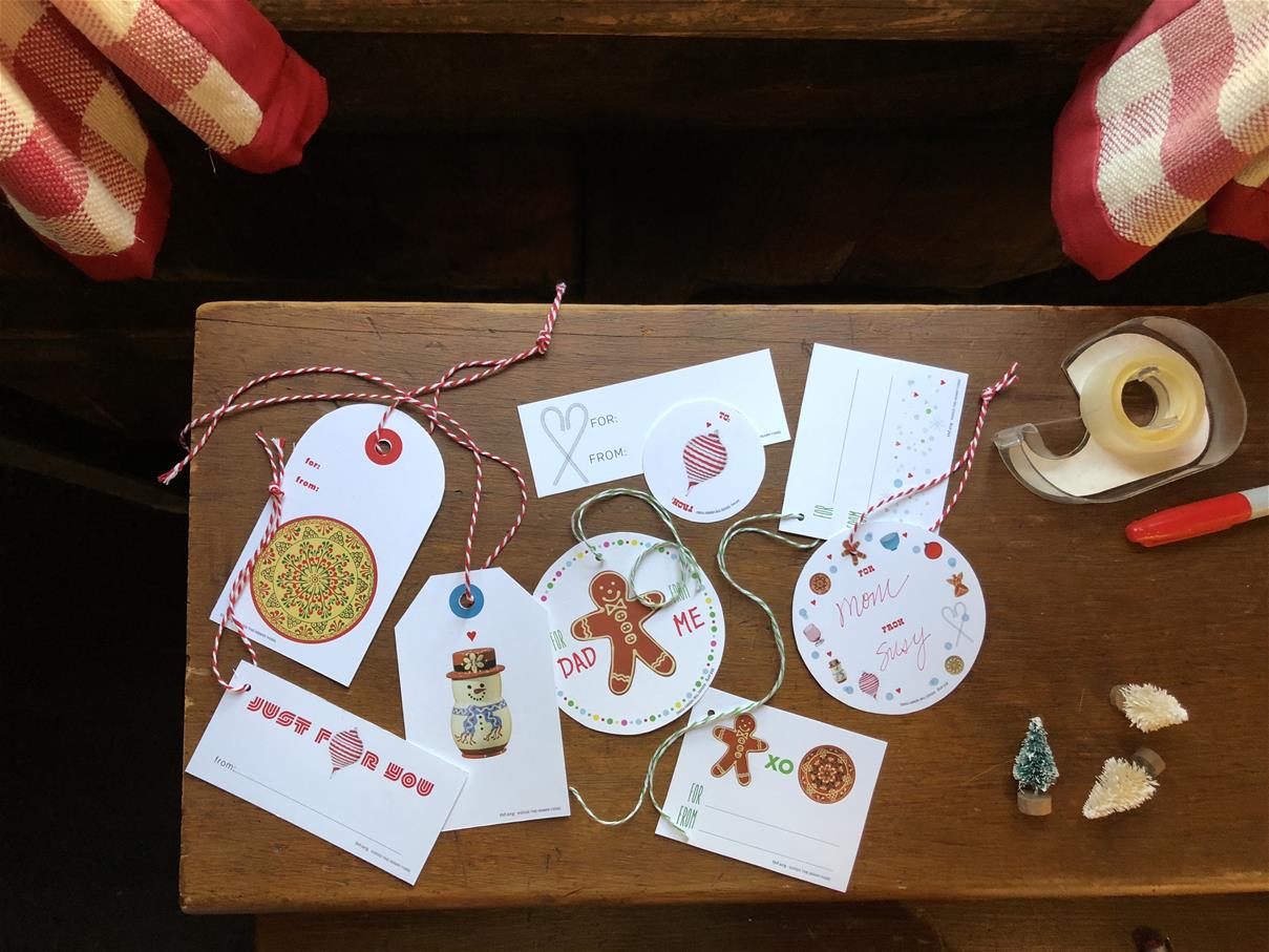 Print-at-Home Holiday Gift Tags Various shapes and designs of holiday labels on a wood table with tape, scissors, and tiny Christmas trees