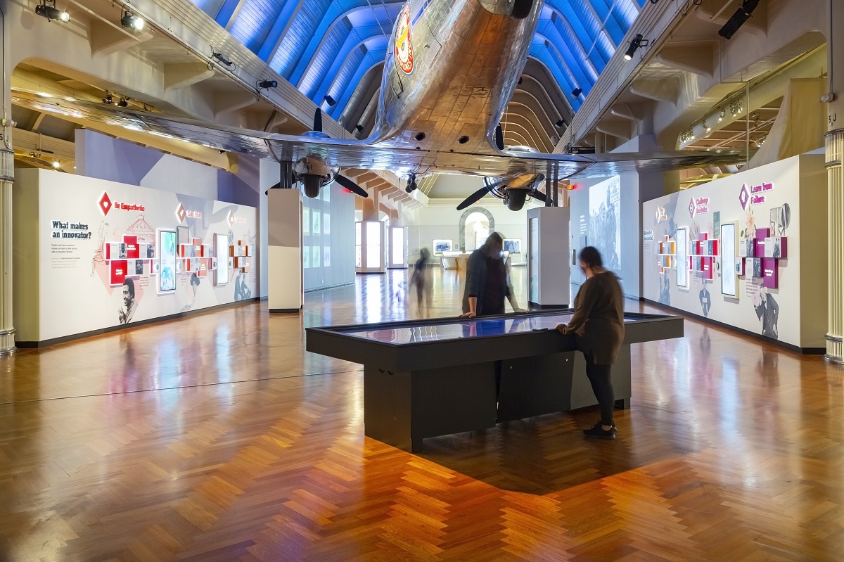 Intersection of Innovation, showing the collections connections table, in Henry Ford Museum of American Innovation Interior space with two people standing at a large table, graphics and text on the walls, and an airplane above