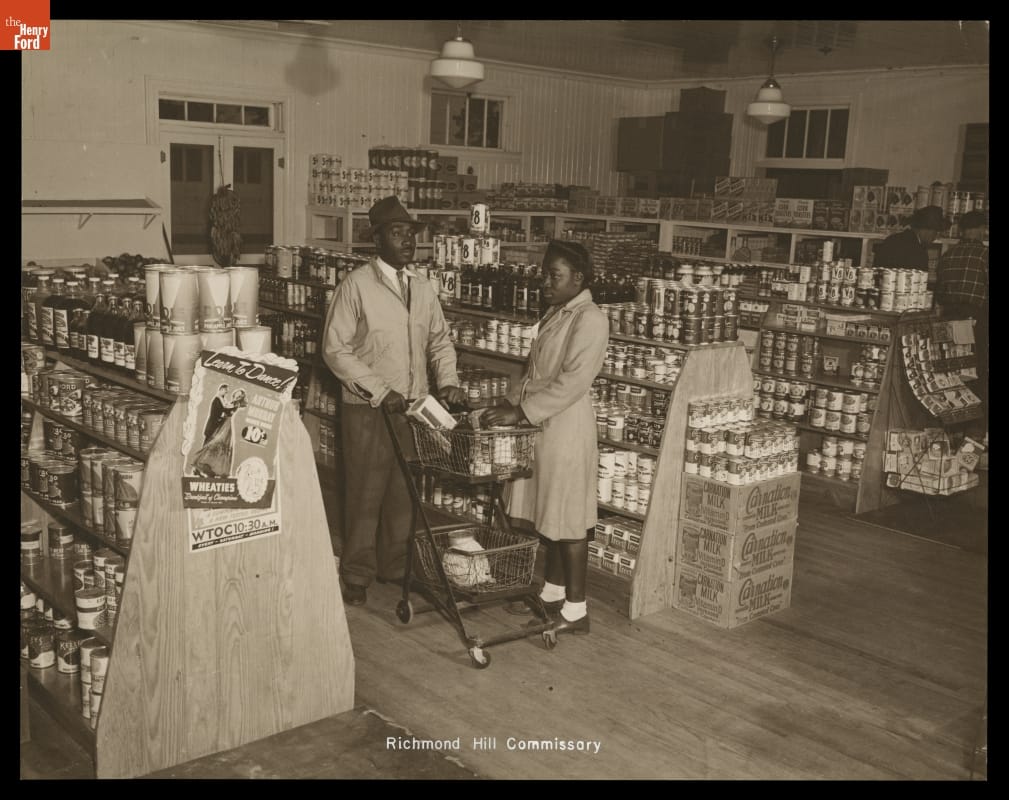Two people stand by a grocery cart in a room filled with shelves stocked with canned goods; other shoppers are nearby