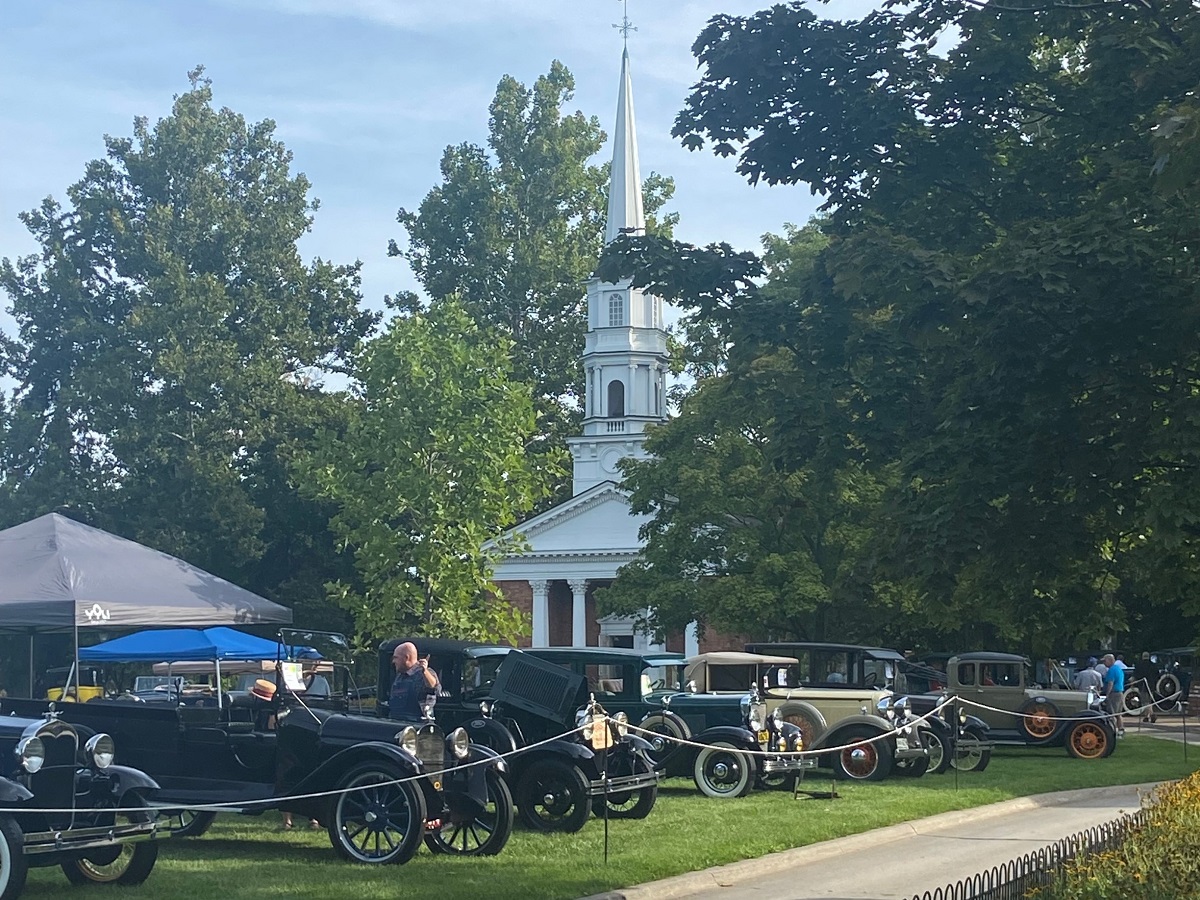 Cars parked on the Village Green in front of Martha-Mary Chapel at Old Car Festival 2022 A row of cars are parked behind a string barrier on a lawn in front of a building with a white wood and red brick steeple; people stand by some of the cars