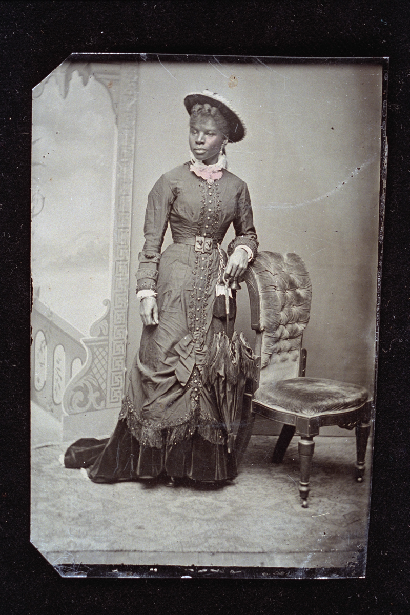 Black woman standing by a chair in front of a backdrop, wearing an elaborate dress and holding a parasol