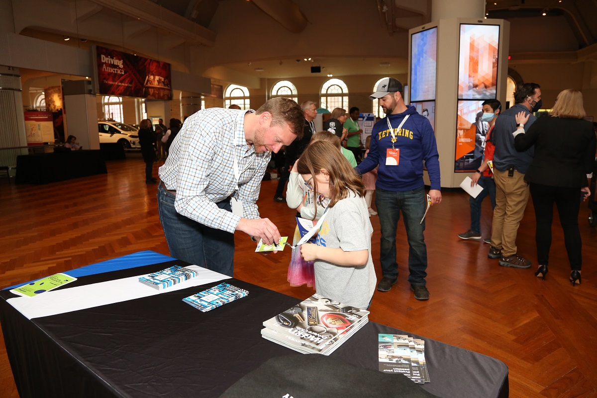 The Hagerty booth at Invention Convention Michigan A man marks a child's lanyard in front of a table in a large space with other people and displays