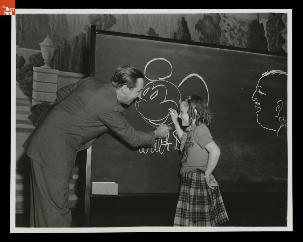 Walt Disney Shows Harriet Bennett How to Draw Mickey Mouse during a Visit to Henry Ford Museum, April 12, 1940 Man in suit and young girl draw Mickey Mouse's head on a chalkboard