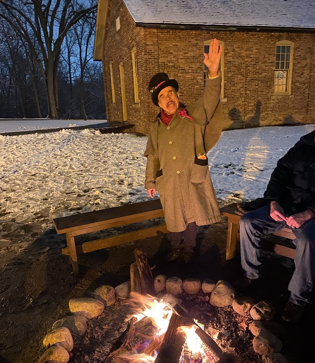 Man in duster coat and top hat by a fire pit waves to the camera