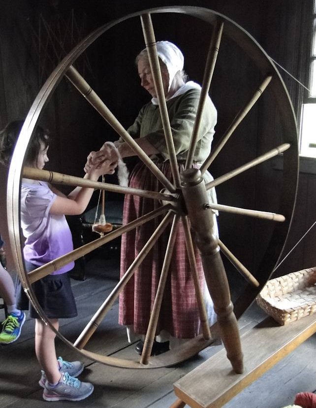 Woman in dress and bonnet hands a spindle to a young girl behind a large spinning wheel 