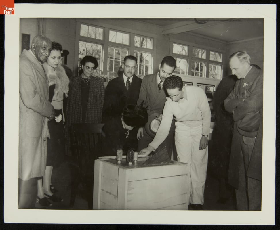 George Washington Carver, Clara Ford, and Henry Ford at Dedication of George Washington Carver Museum, March 1941 Person wearing hat sits, head tipped down toward a wooden crate, while other people look on