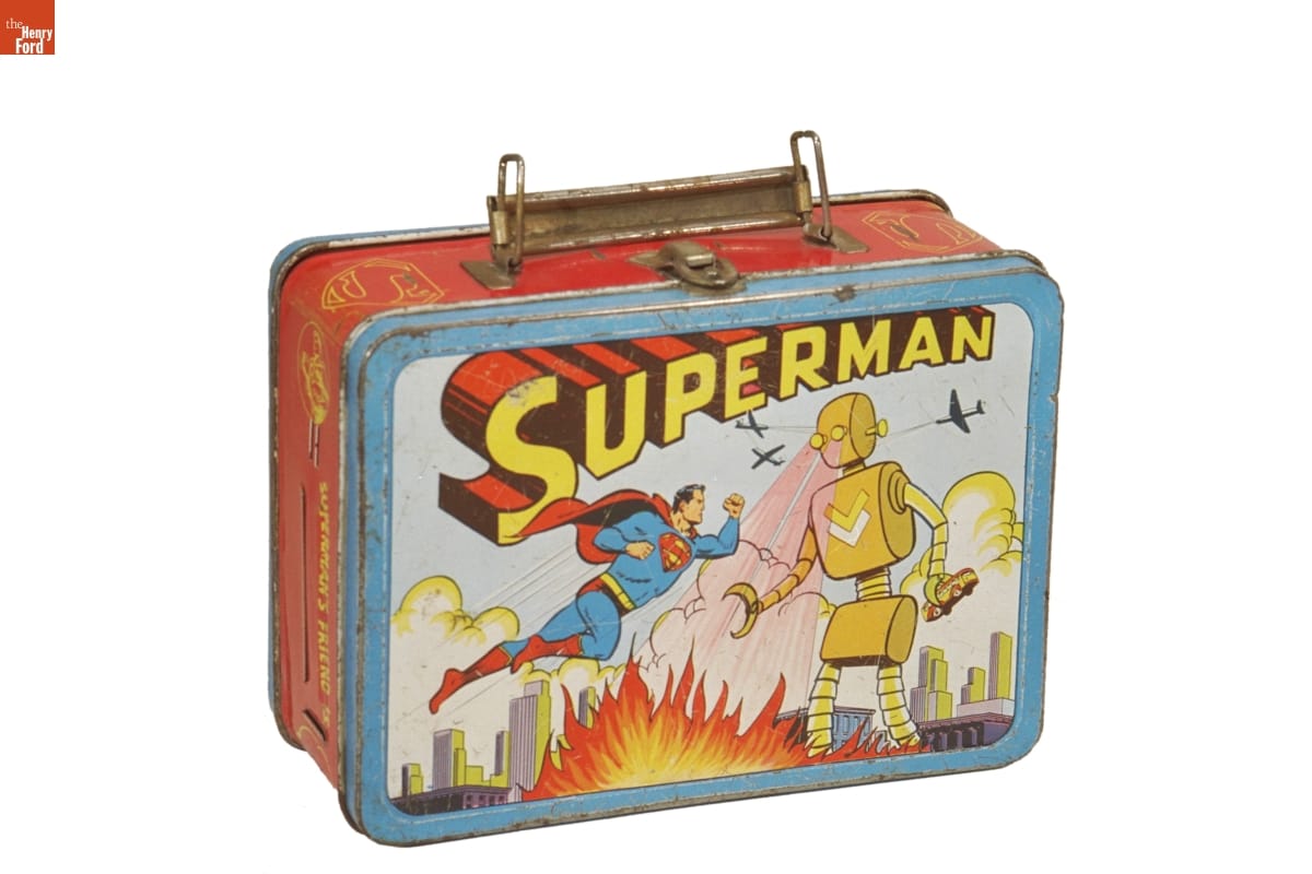 Slightly rusted lunchbox with handle and front panel illustration of man in blue and red super hero outfit floating in air and fighting a large yellow robot shooting flames from its eyes; 3D text reading “Superman”