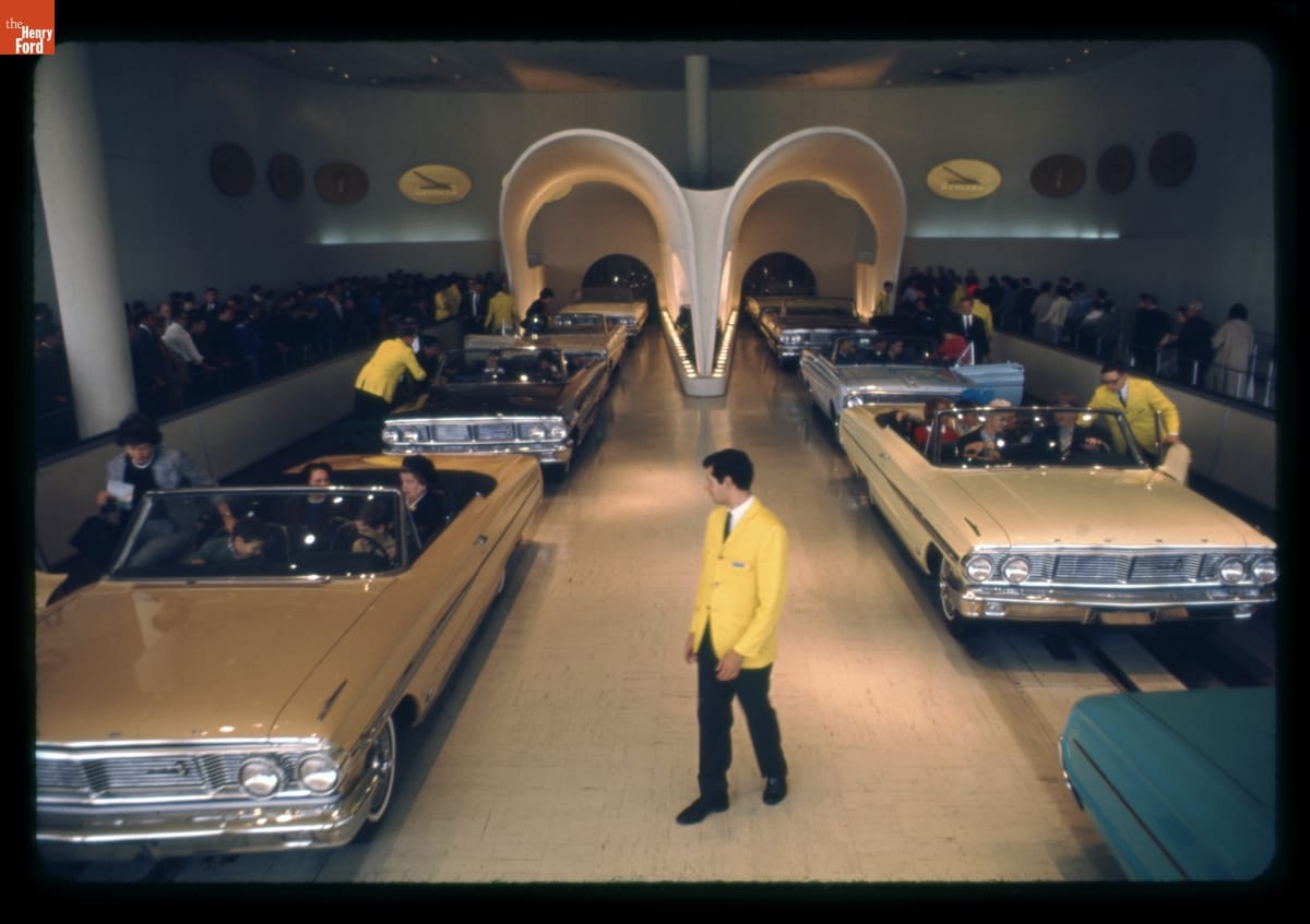 Loading Area for the Magic Skyway Ride at the Ford Pavilion, New York World's Fair, 1964-1965 Assistants in yellow sports jackets assist people into convertible cars on two sets of tracks in a large room; more people queue on either side, waiting their turn