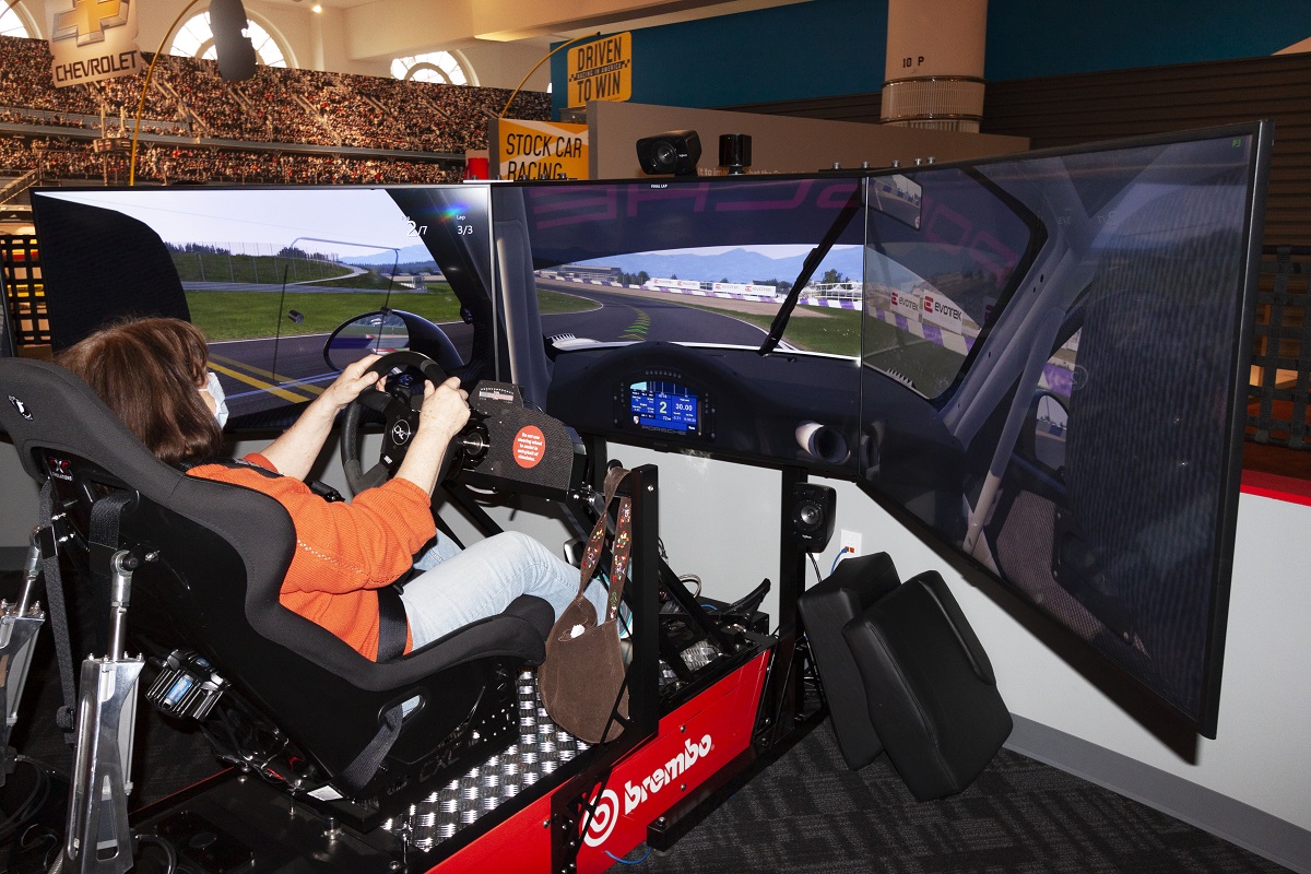 A guest experiences racing in the new Driven to Win exhibit A person sits in a car simulator in front of three large screens displaying a race track