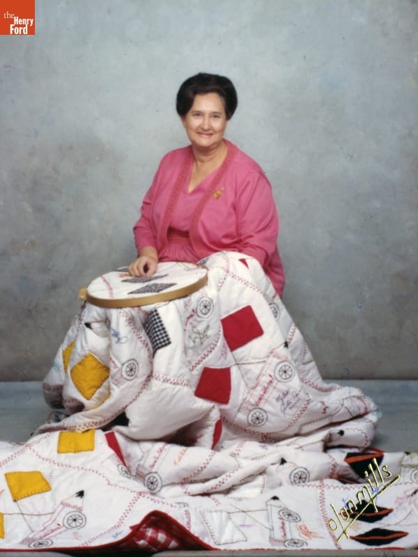 Jeanetta Holder with Her Indianapolis 500 Quilt Made for Bobby Unser, 1975-1980 Woman with pink outfit and dark hair sits with a quilt over her, part of which is in a needlework frame on her lap
