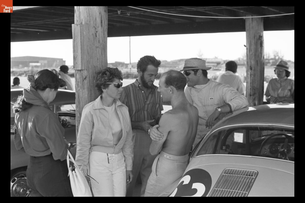 Denise McCluggage Talking with Stirling Moss at Bahamas Speed Weeks, November 27 - December 10, 1961 Group of people stand and talk under a pier or bridge surrounded by cars