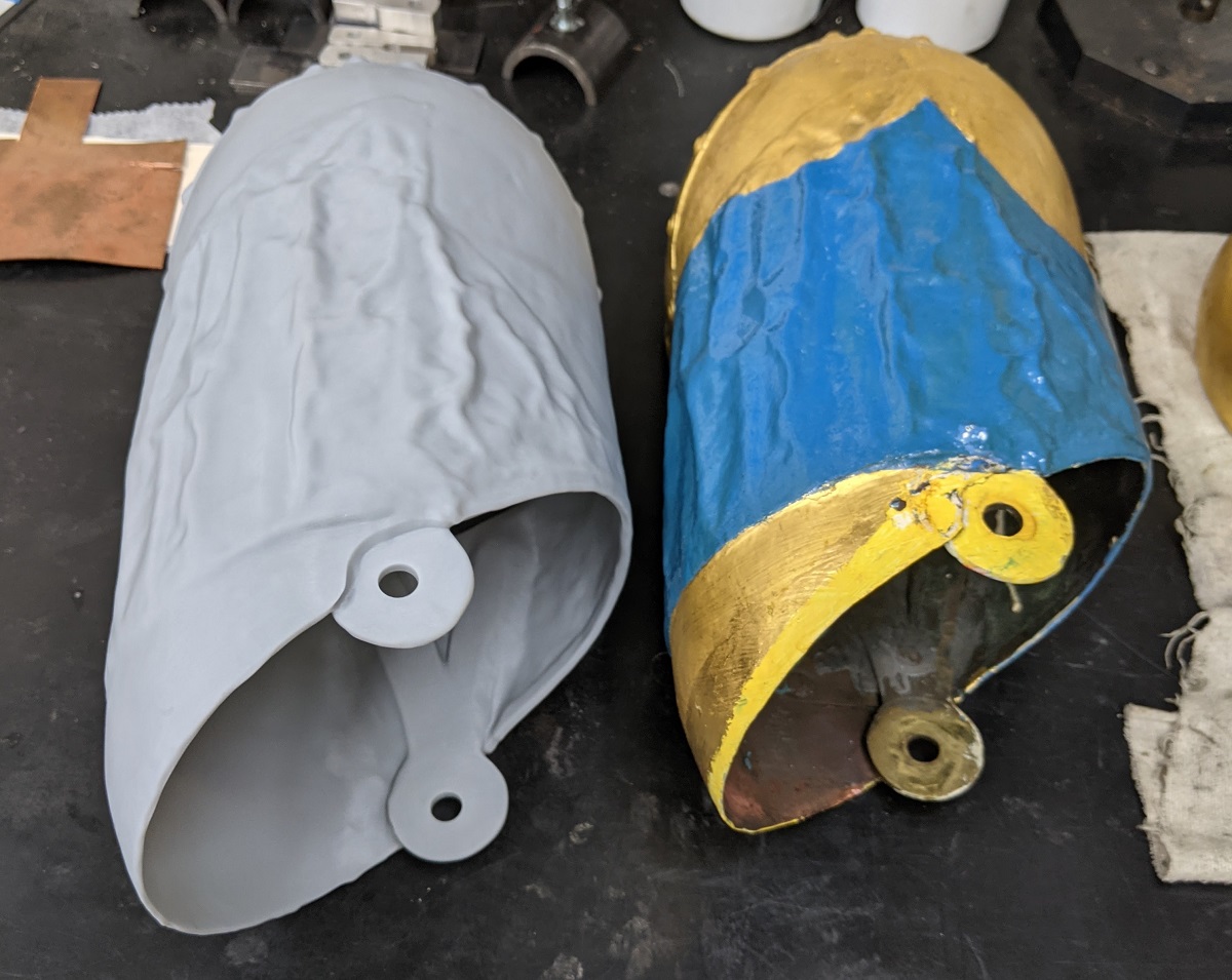 A side-by-side comparison between the SLA 3D-printed copy on the left and the original artifact on the right Two hollow shapes--one gray, one painted yellow and blue, sitting on a workbench