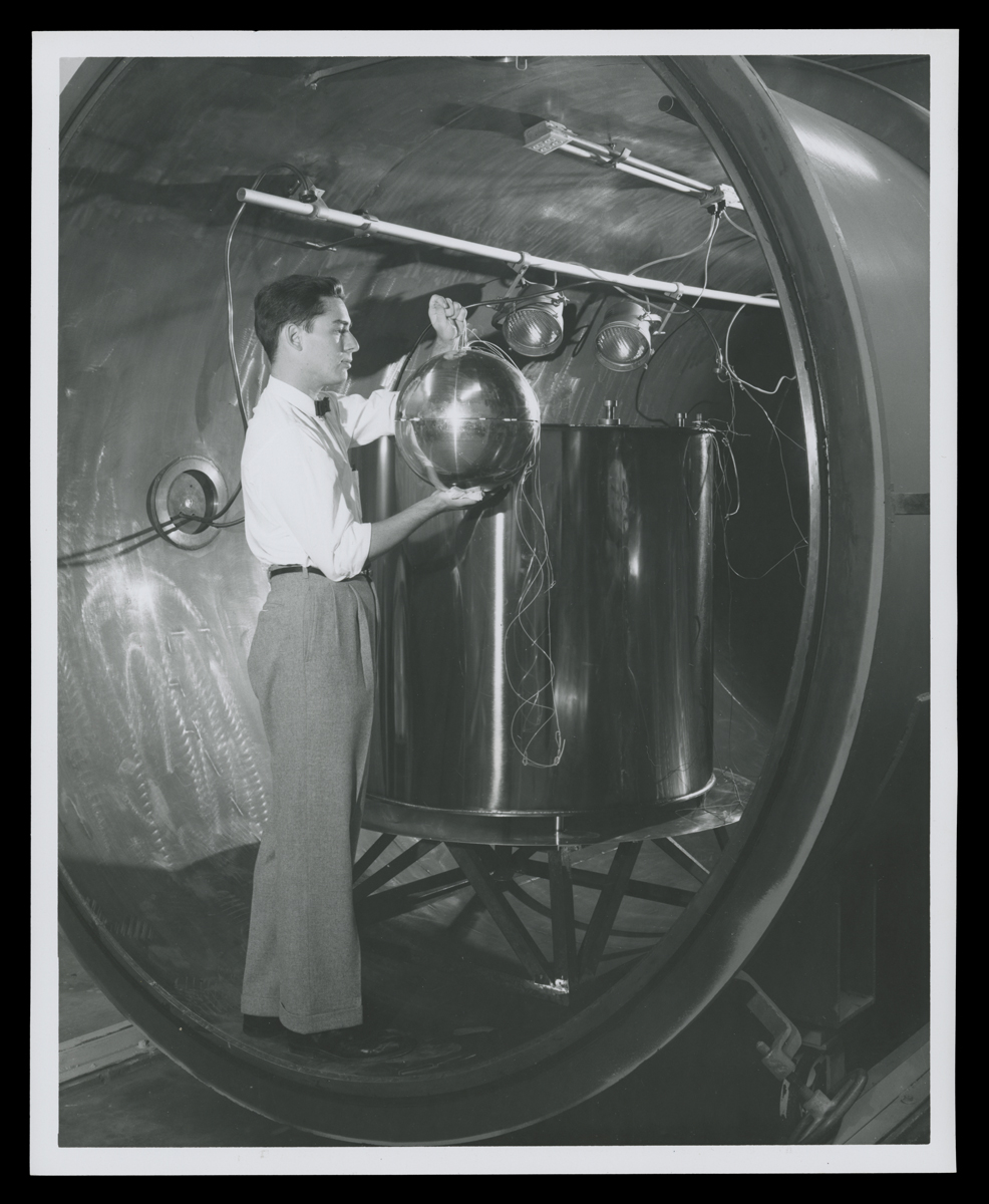 Testing the Lunar Capsule Built by Ford Motor Company Aeronutronic Division, 1960 Man stands in large metal tube holding shiny sphere next to large vat