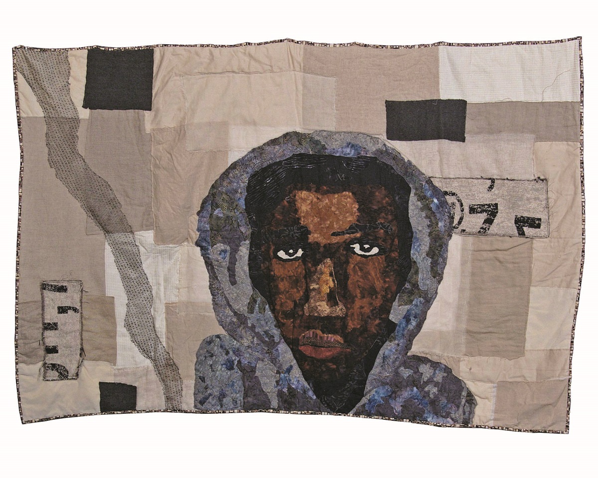 Sara Trail’s handmade quilt, Rest in Power, commemorates the life of Trayvon Martin Quilt with background of black and gray blocks of various sizes and face and shoulders of dark-skinned man in a gray hoodie