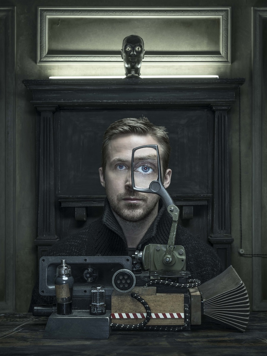 Ryan Gosling, photographed by Dan Winters Man sits behind machine with magnifying glass in front of one eye, enlarging it, in front of architectural details and lit-up doll's head