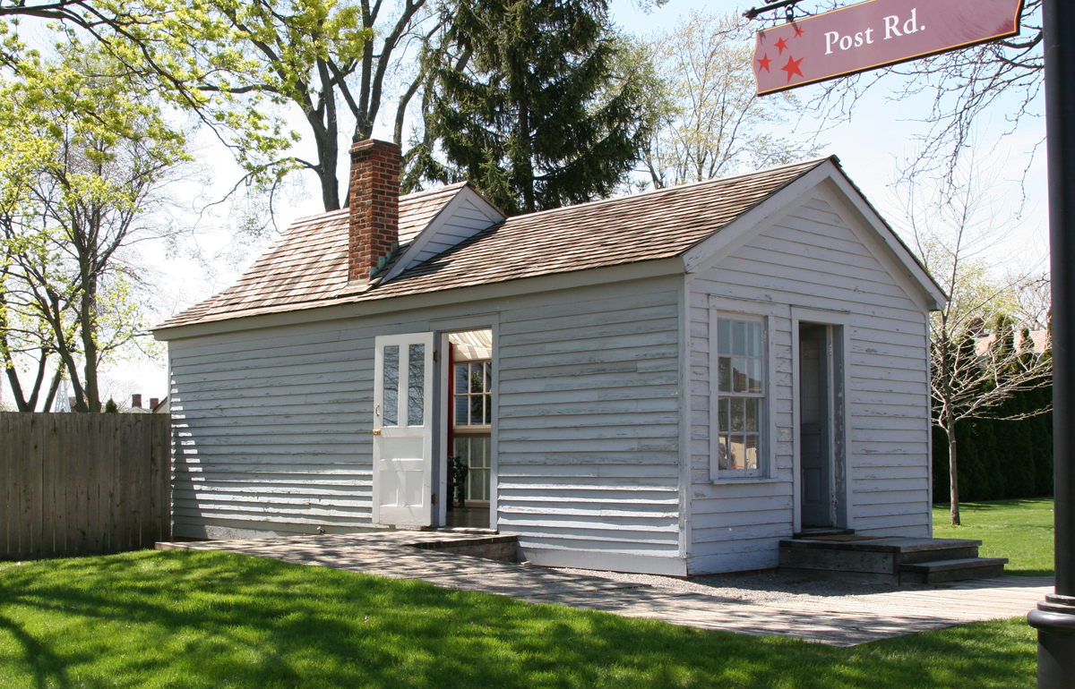 Small wood building with doors in front and side, one window in the front, and a chimney