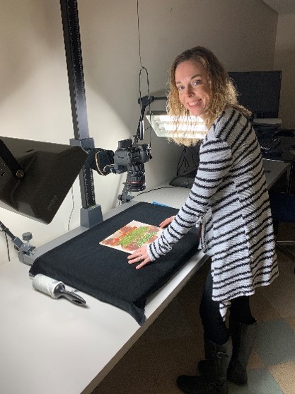 Project Digitization Specialist Karen Wissink Images an Artifact. Woman looking at camera, standing at table setup with lights and camera