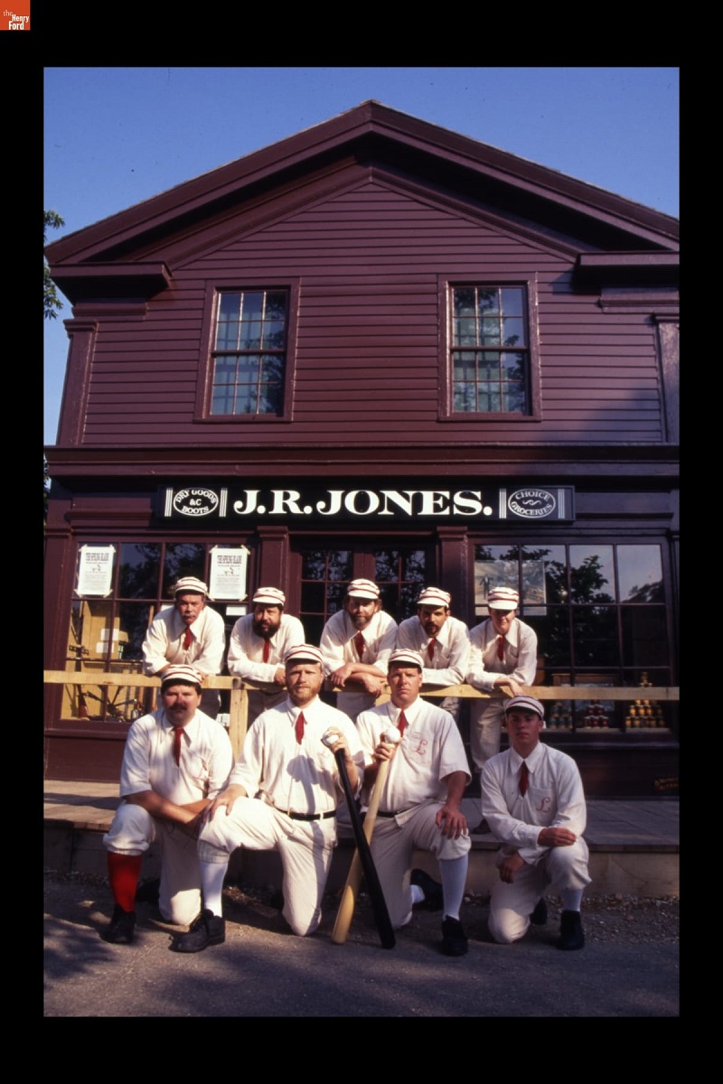 "Lah-De-Dahs" Baseball Team in Greenfield Village, Spring 1994 Nine people in old-fashioned baseball uniforms stand and kneel in front of a maroon wooden building with sign "J.R. Jones"