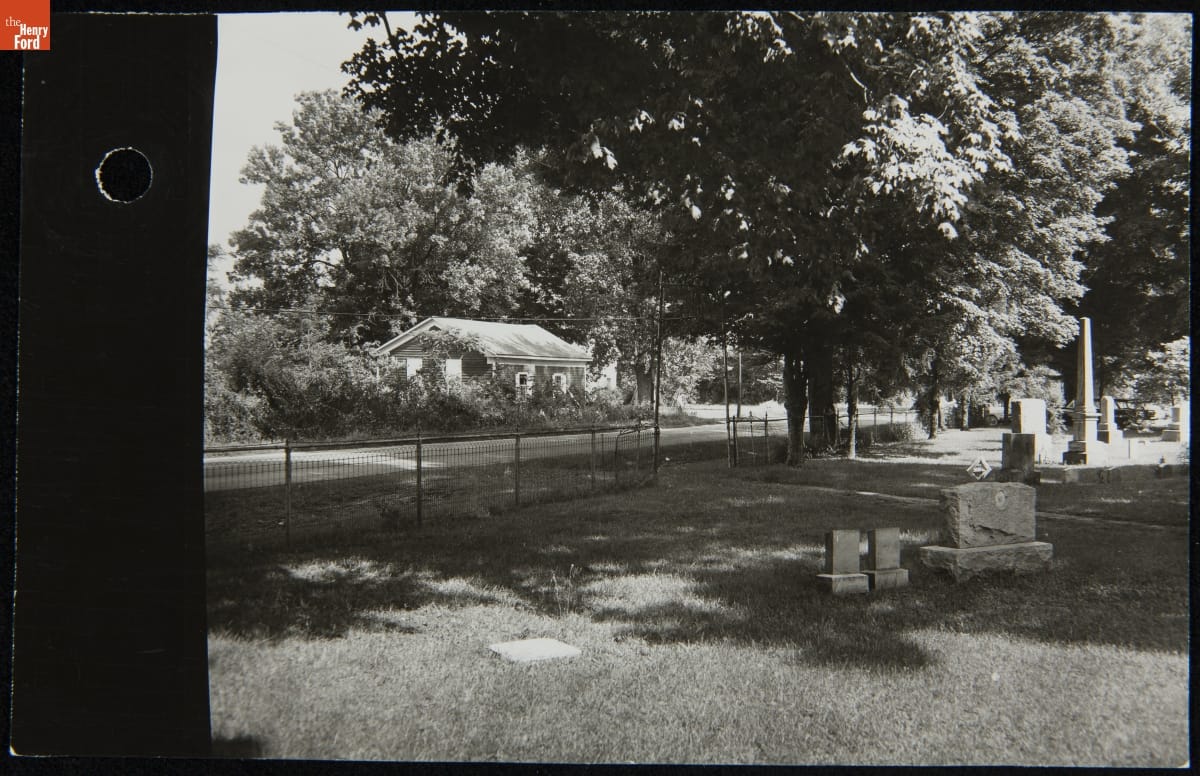 View of Dr. Howard's Office from the Cemetery across the Street, Tekonsha, Michigan, August 1959 Black-and-white photo of cemetery, road, and small, overgrown building across the road