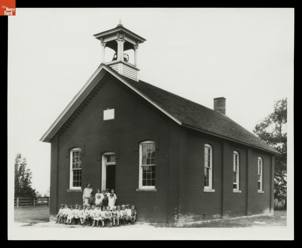 Woman poses for photo with a group of young children outside a small brick building