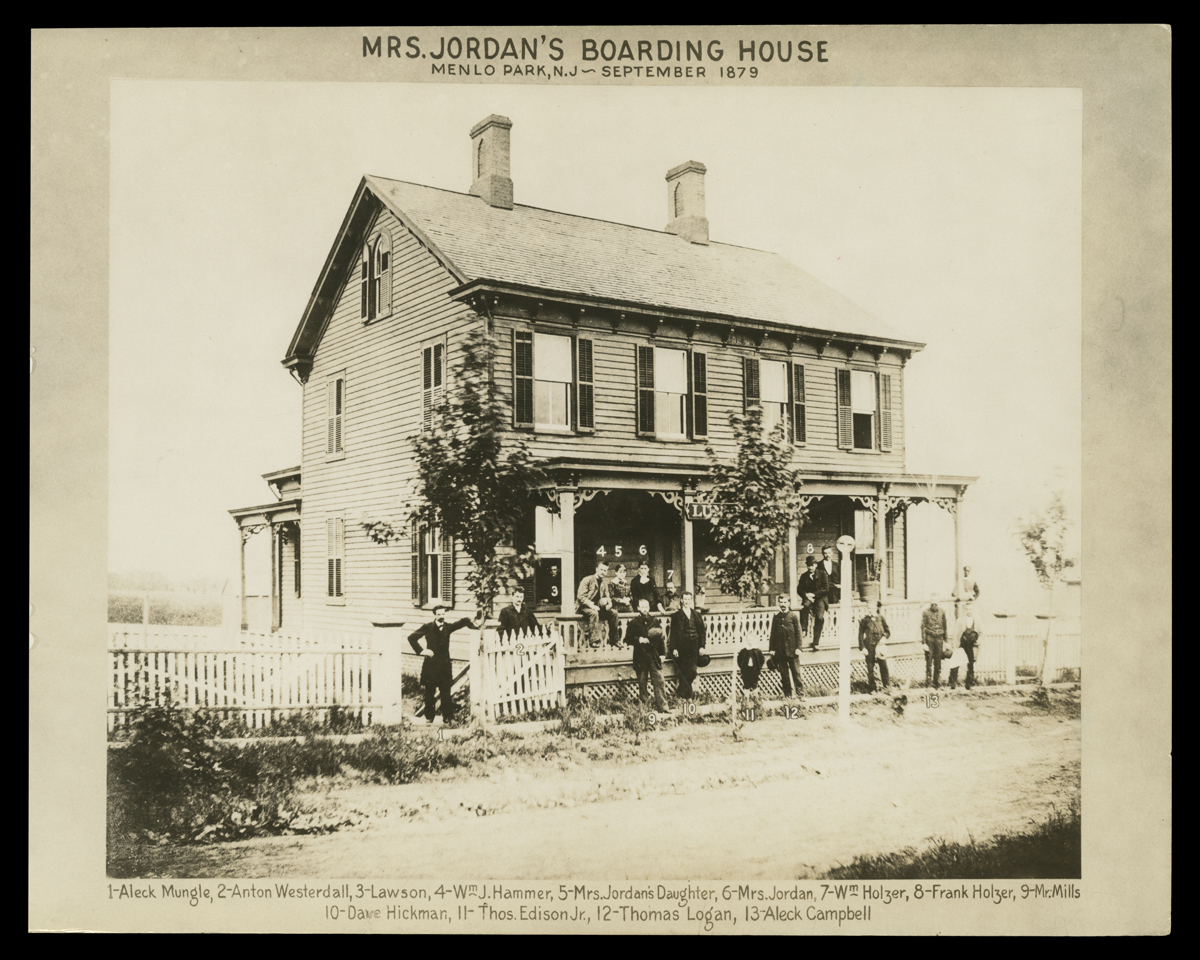 Black-and-white photo of two-story wooden house with people on porch and standing by and in front; also contains text