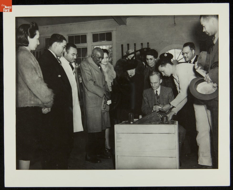 Henry Ford Inscribing Cement Block at Dedication of George Washington Carver Museum, March 1941 Man sits at crate while other people look on