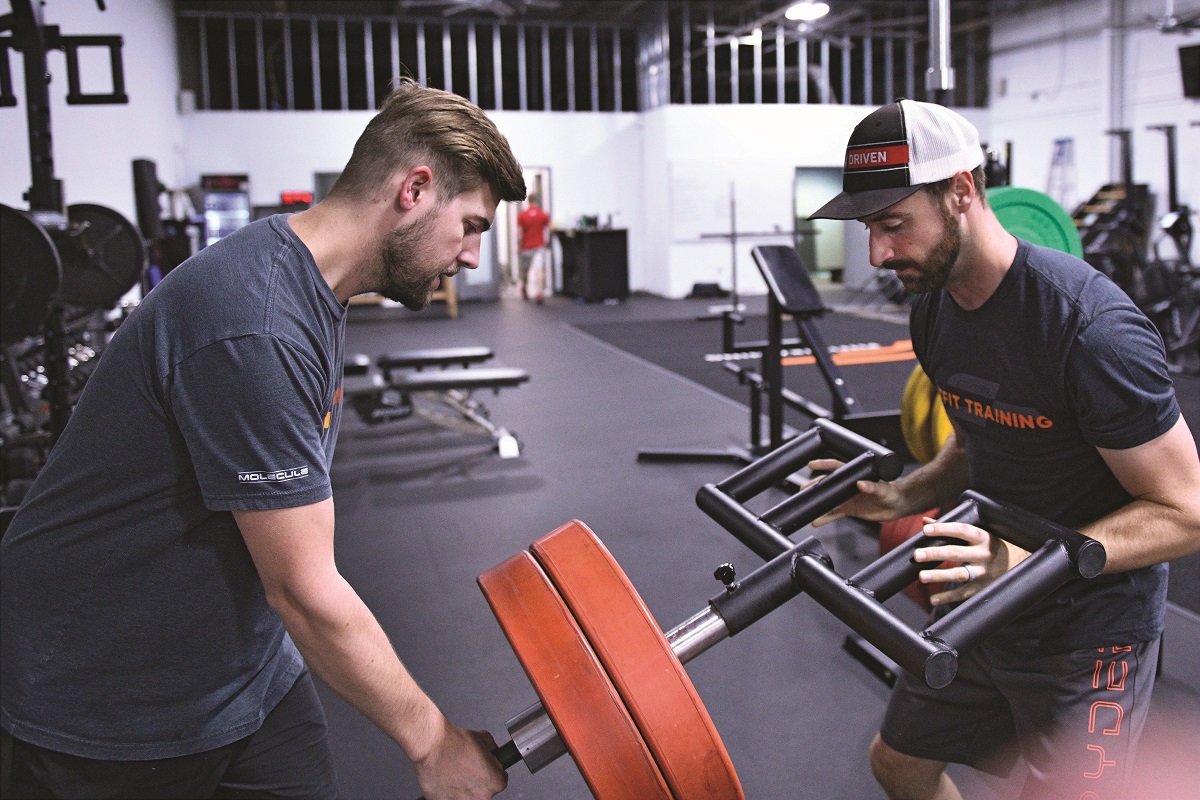 Driver James Hinchcliffe strength training at PitFit Two men in t-shirts stand on either side of a fitness machine in a gym