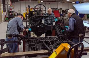 Attaching the cultivator rear track sweep attachment Three men wearing blue gloves work on a piece of equipment
