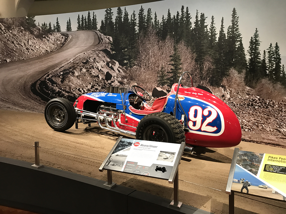 The 1958 Moore/Unser racing up our scenic recreation of Pikes Peak in Driven to Win. Red and blue race car with large number "92" on side sits on a dirt-like surface in front of a backdrop of a mountain road and trees; placards in front