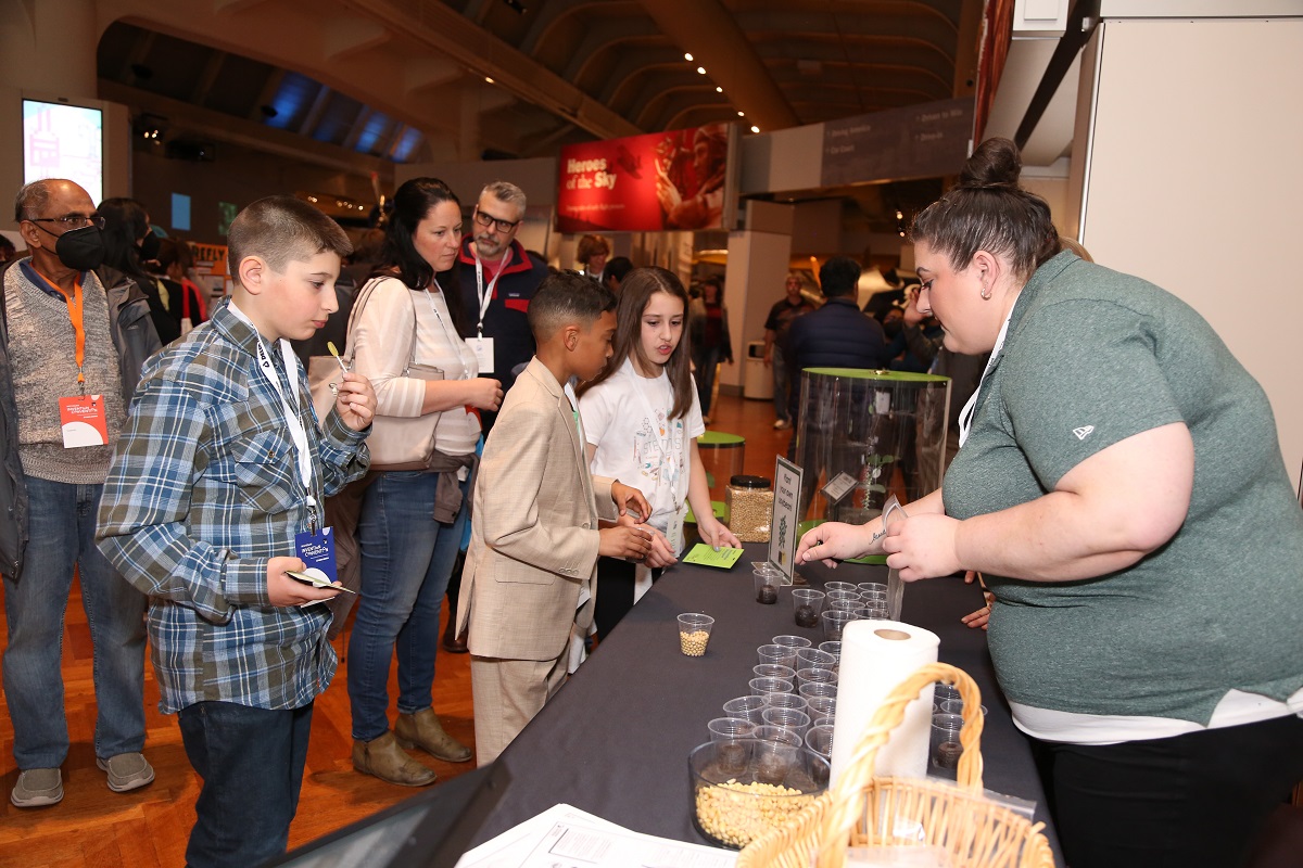 The Michigan Soybean Committee booth at Invention Convention Michigan A group of children and adults stand by a table filled with small cups, behind which is a woman in a gray shirt