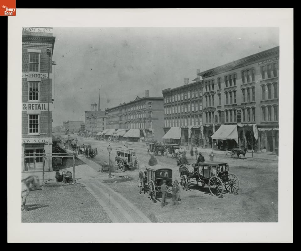 City street scene with buildings, horse-drawn carriages, and pedestrians