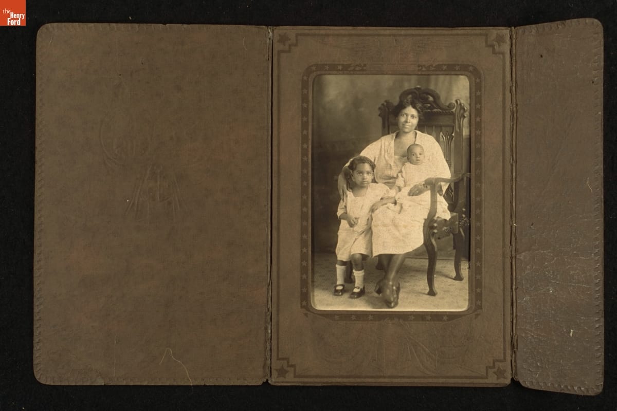 Black woman, seated, with infant on lap and toddler standing beside her