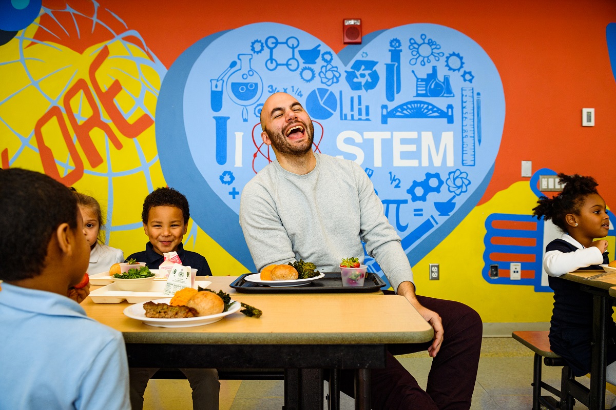 Dan Giusti with students at Winthrop Elementary School A man, laughing, sits at a table with children, all with trays of food, in front of a brightly-painted wall