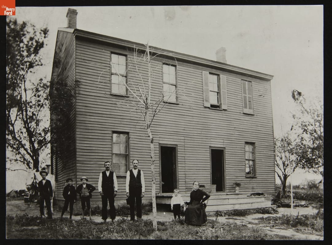 Seven children and adults stand and sit outside a two-story wooden building